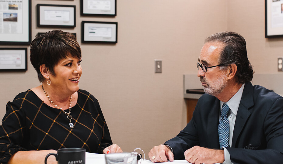 Two people in business attire sit at a table in an office, engaged in conversation, with documents, a pen, and a coffee mug in front of them. Framed certificates hang on the wall.