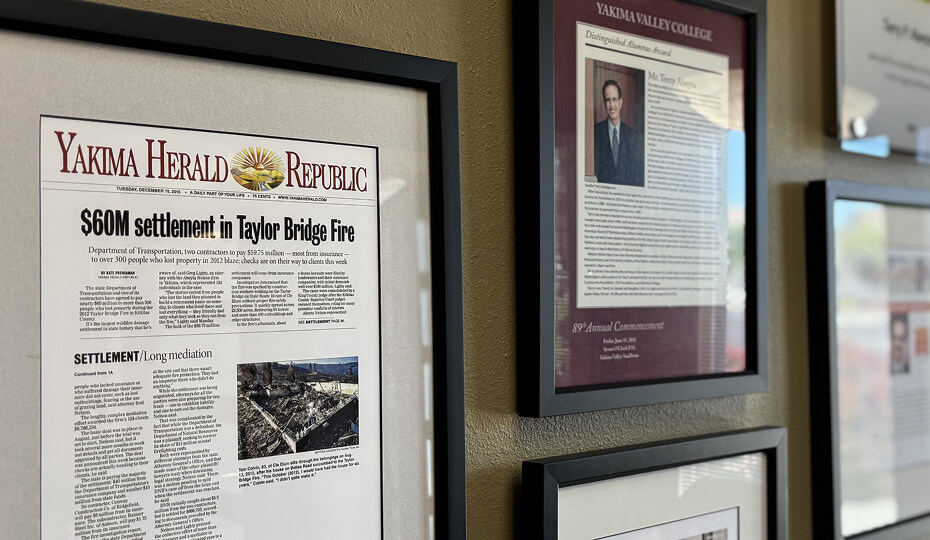Framed newspaper article about a $60M settlement in the Taylor Bridge Fire and several plaques hang on a beige wall.