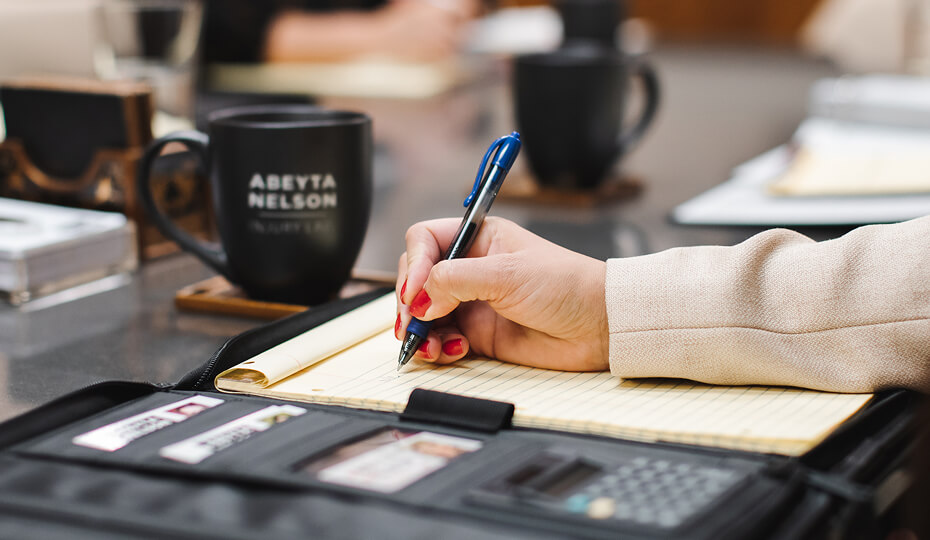 A person writes on a yellow legal pad with a pen at a conference table; coffee mugs and office supplies are visible nearby.