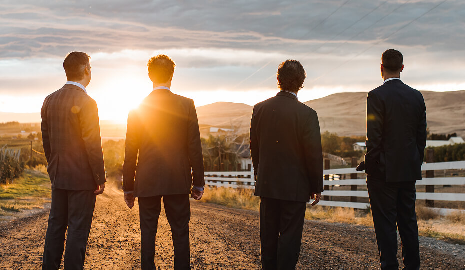 Four men in suits stand on a dirt road facing a sunset, with hills and a white fence in the background.