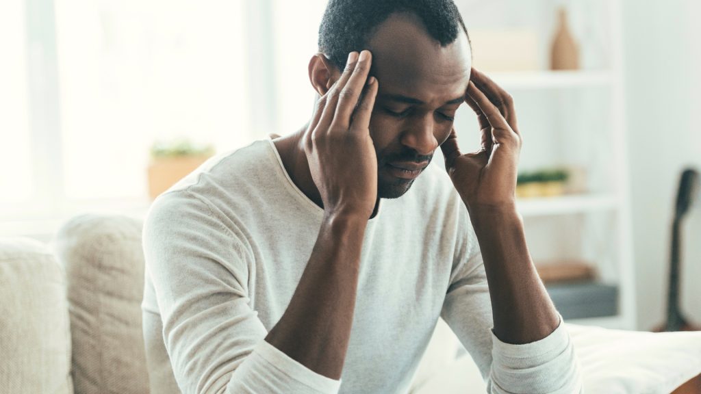 A man sitting on a couch holds his temples with both hands and appears to be experiencing a headache or stress.