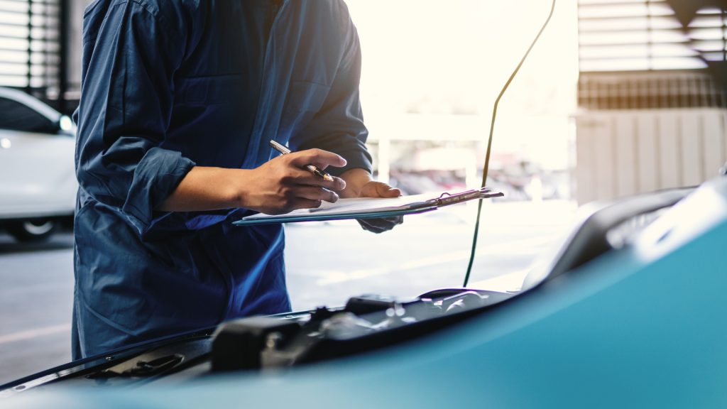 A mechanic wearing a blue uniform writes on a clipboard while inspecting a car in an auto repair shop.