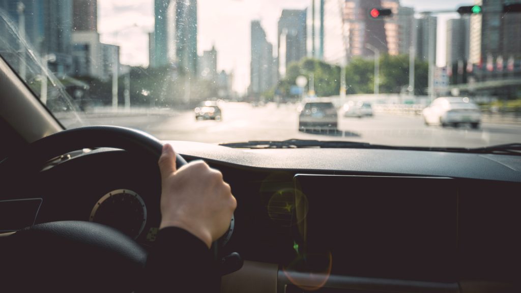 Person driving a car through a city, view from inside the vehicle showing hands on the steering wheel and blurred buildings ahead.