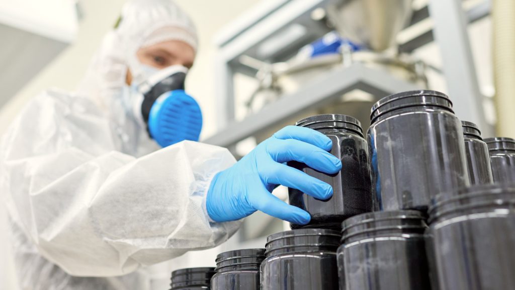 Person in protective suit and gloves handling black plastic jars in an industrial or laboratory setting.