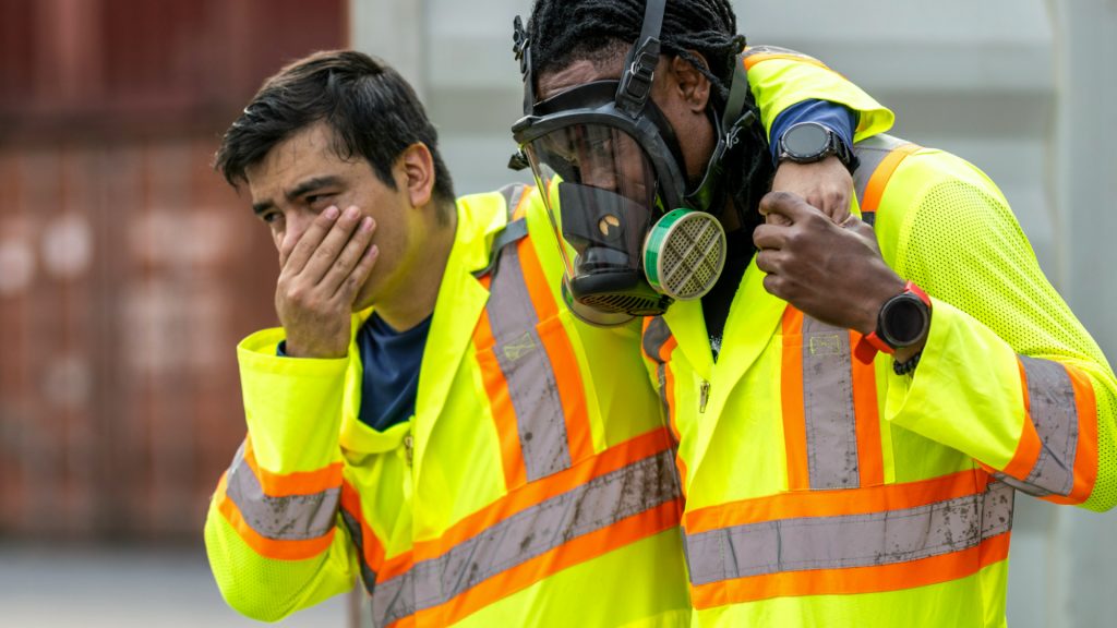Two workers in high-visibility jackets; one wears a gas mask and supports the other, who covers his nose and mouth, suggesting exposure to hazardous fumes.