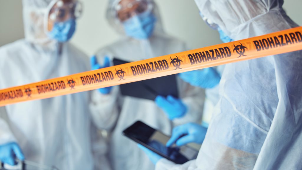 People in protective suits and masks working behind a yellow biohazard tape, with some holding electronic tablets in a controlled environment.