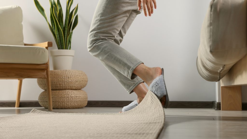 Person tripping over the edge of a raised rug in a living room with a plant and woven stools in the background.