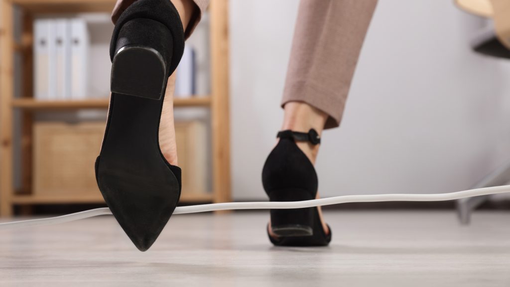 A person in black heels and beige pants is about to trip over a white electrical cord on the floor in an office setting.