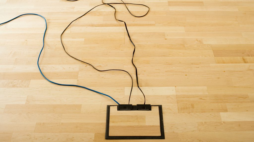 Two cables, one blue and one black, are plugged into two devices placed in a black rectangular frame on a light wood floor.