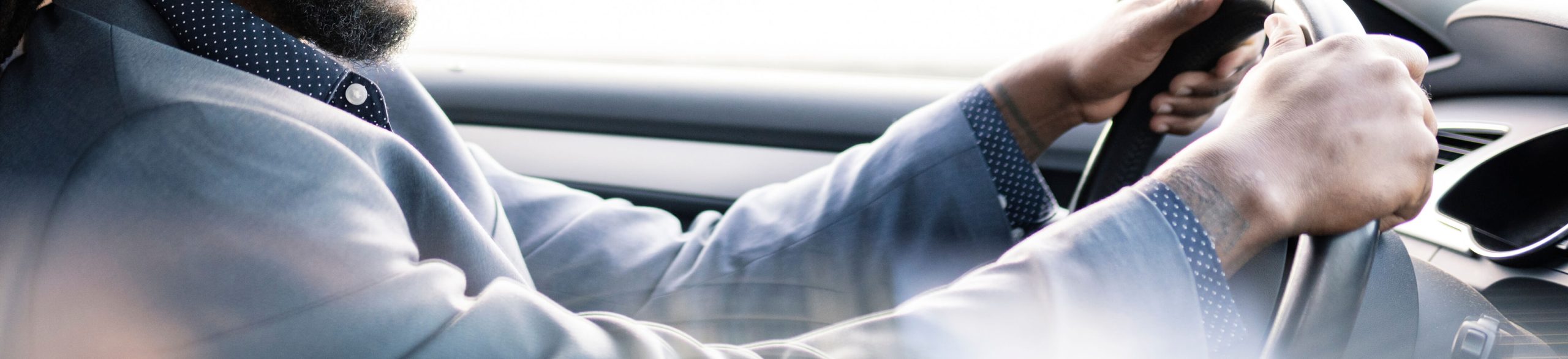 Una persona vestida con un traje y una camisa de lunares conduce un coche, sujetando el volante con ambas manos.