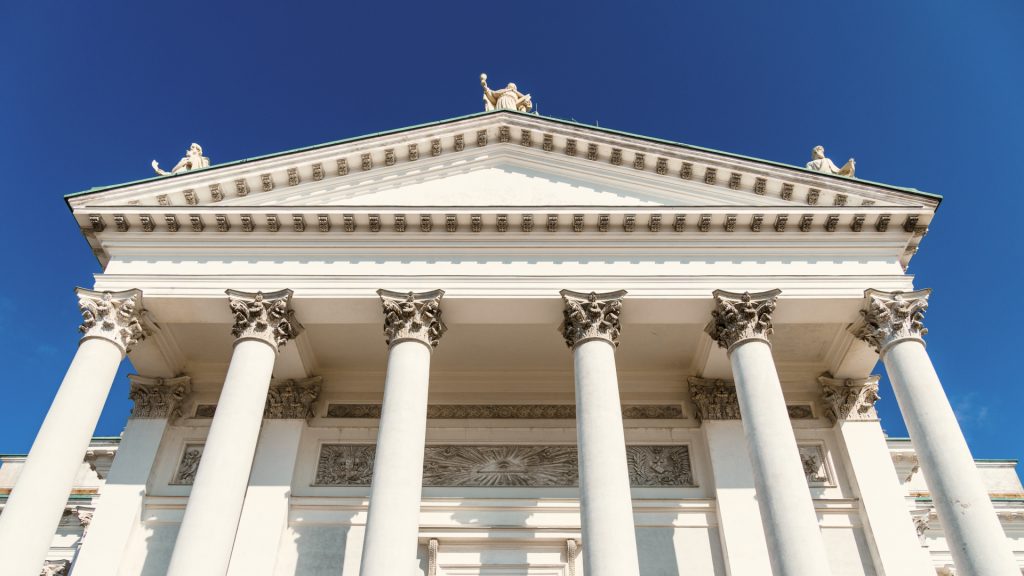 Edificio neoclásico blanco con columnas altas, capiteles decorativos y estatuas en el tejado, visto desde abajo contra un cielo azul despejado.
