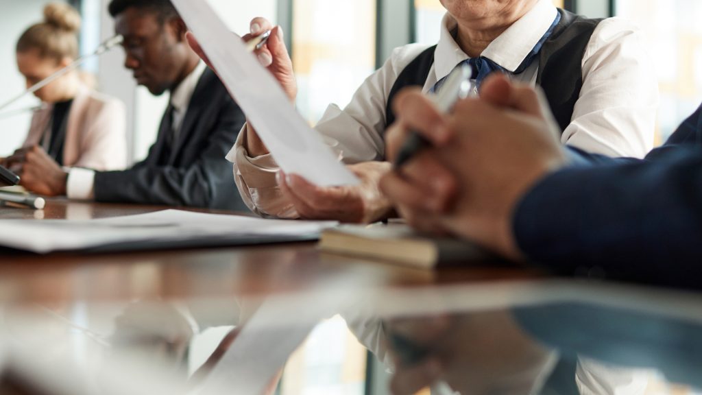 Personas con vestimenta de negocios sentadas en una mesa de conferencias con documentos, bolígrafos y cuadernos, participando en una reunión o discusión.