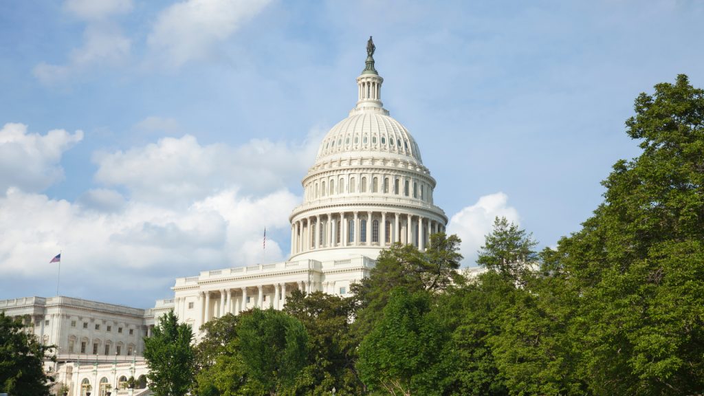 El edificio del Capitolio de los Estados Unidos con su cúpula y estatua en la parte superior, parcialmente oculto por árboles, bajo un cielo parcialmente nublado.