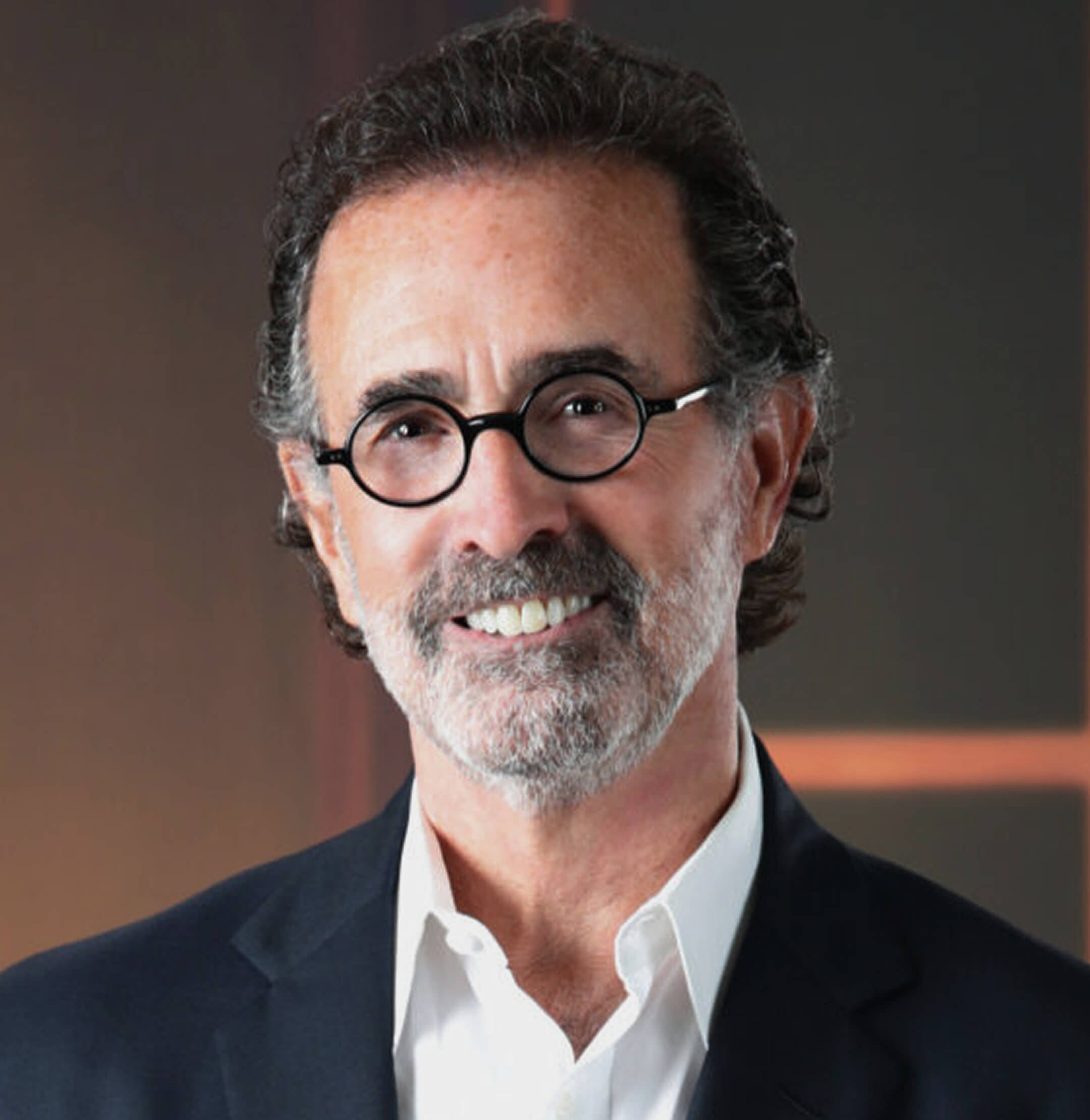 A middle-aged man with curly dark hair, a beard, and round glasses, wearing a dark suit jacket and white shirt, smiling in front of a blurred background.
