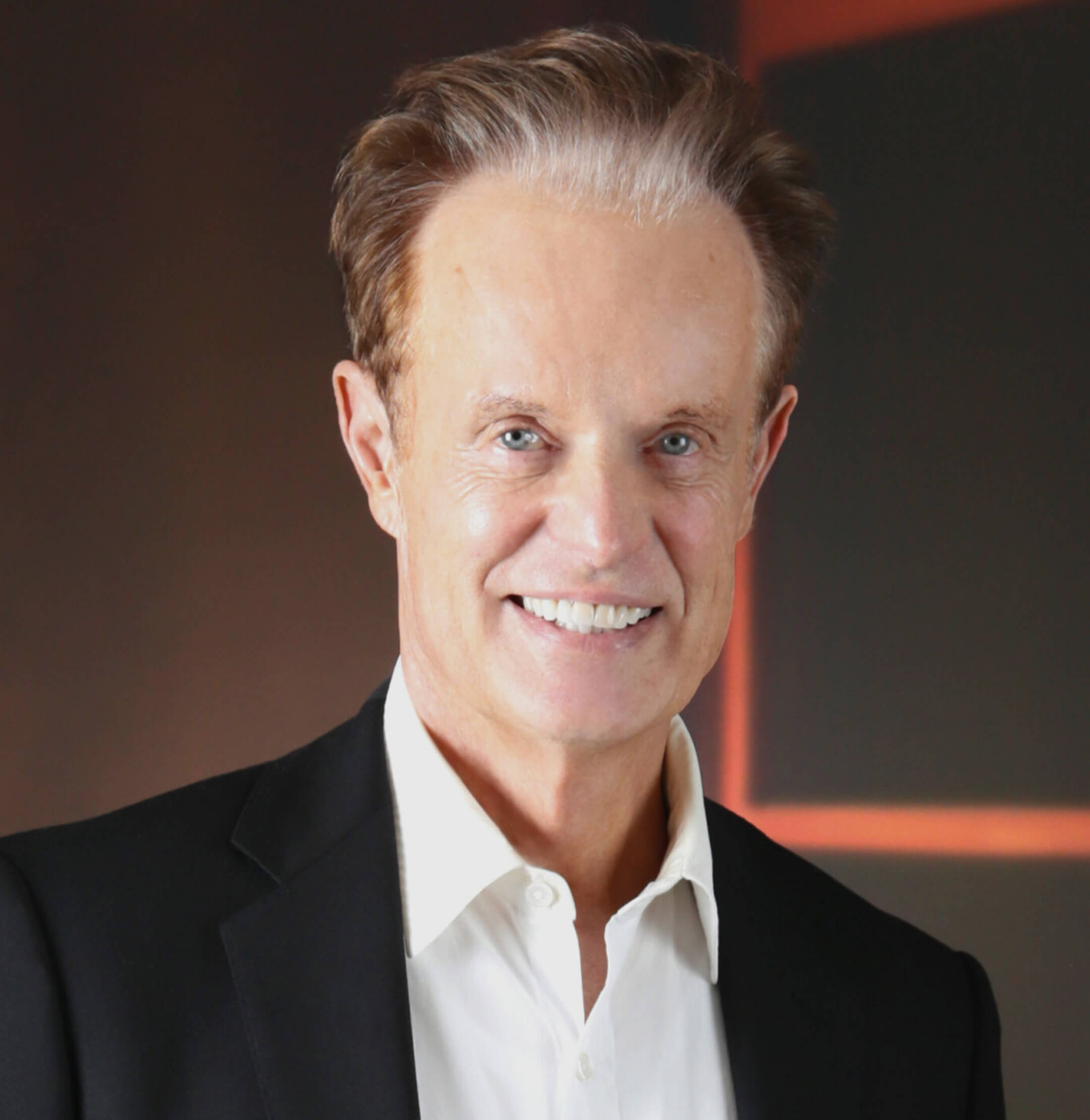 A man with light skin and short brown hair, wearing a white dress shirt and black blazer, smiling at the camera with a blurred dark background.