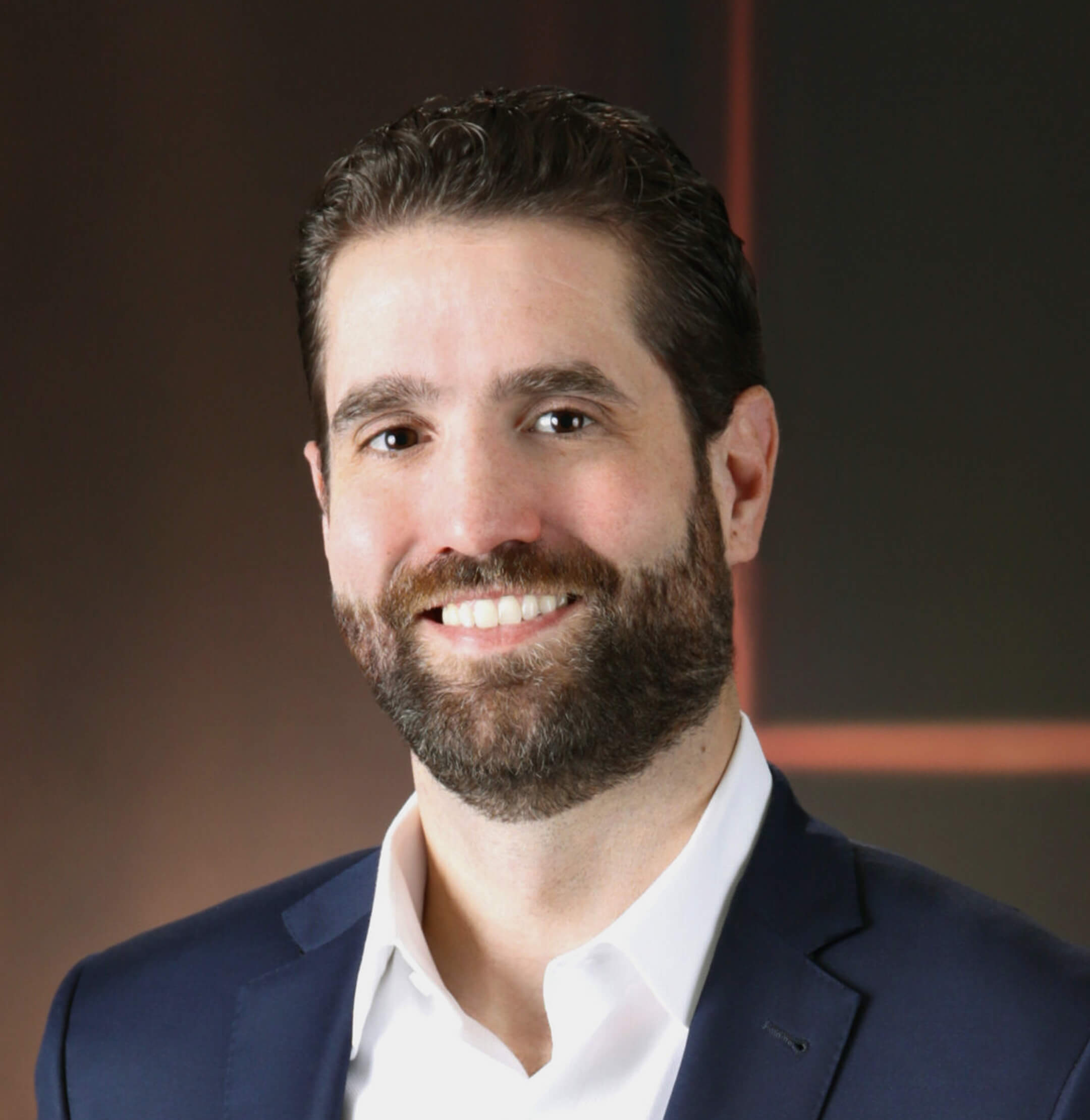 A man with dark hair and a beard, wearing a navy suit and white shirt, smiling in front of a dark background with orange lines.