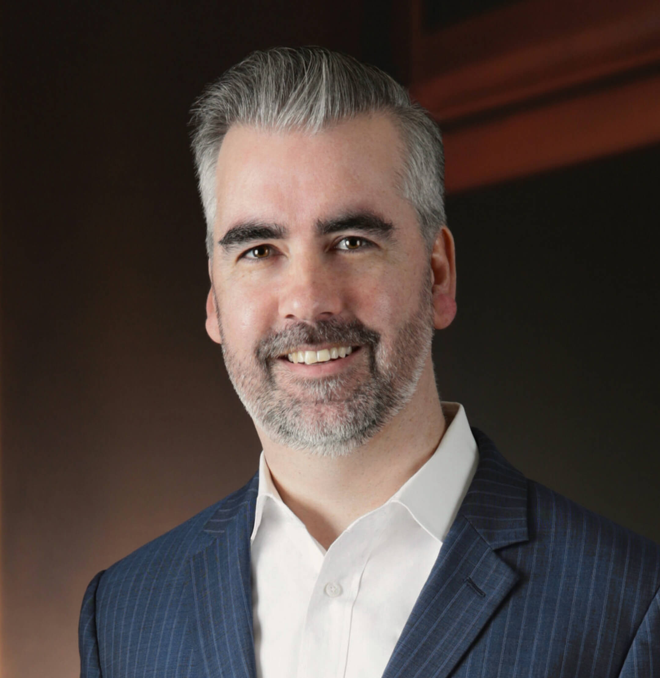 A man with short gray hair and beard wearing a white shirt and navy pinstripe suit jacket, smiling at the camera against a dark background.