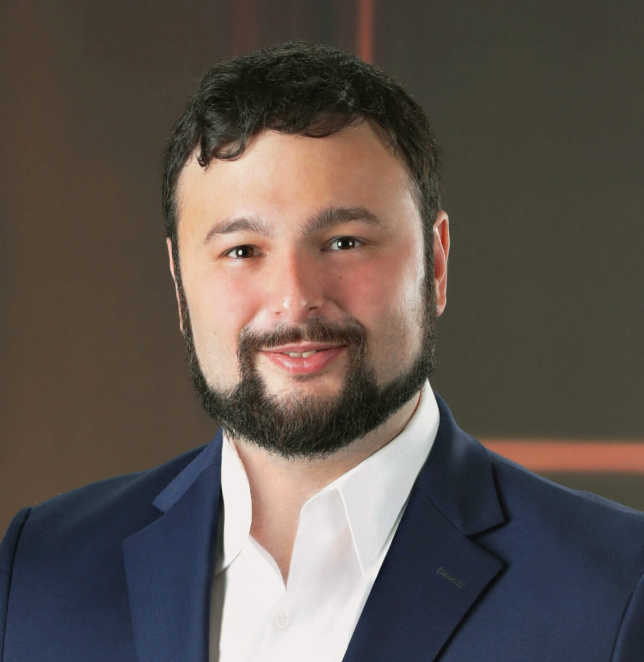 A man with dark hair and a beard wears a navy blue suit jacket and white shirt, posing in front of a plain, dark background.