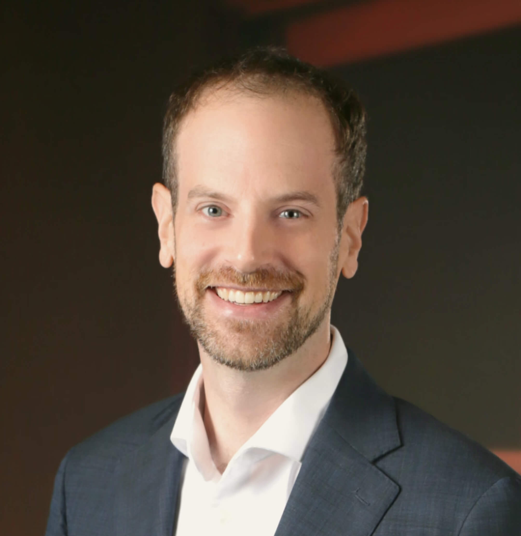 A man with short brown hair and a beard, wearing a navy suit jacket and white shirt, smiling in front of a dark background with red accents.