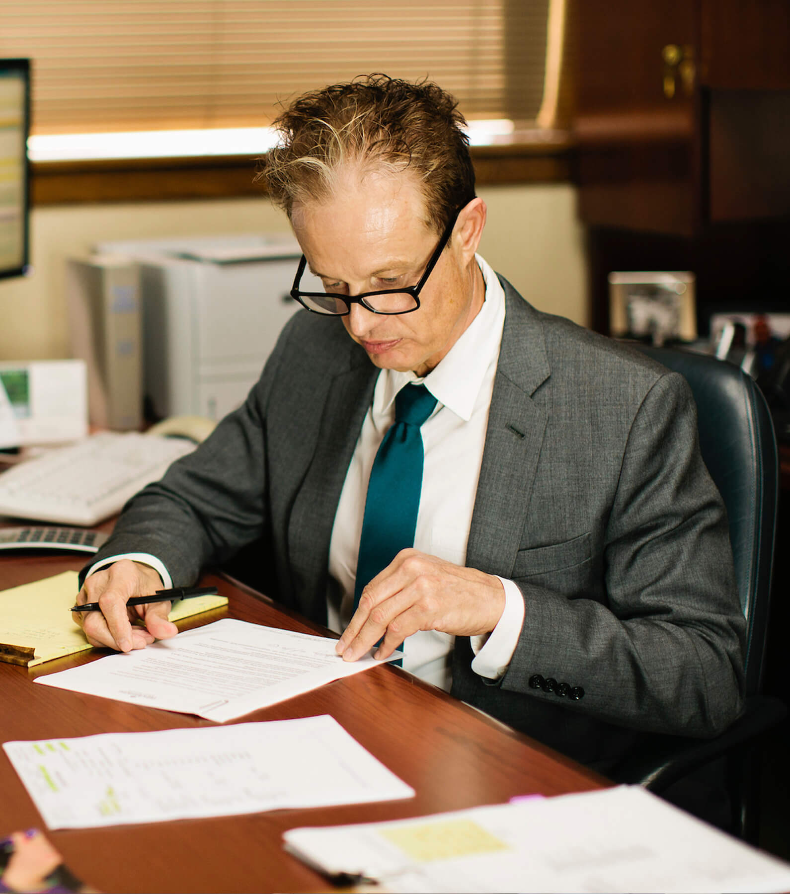 A man in a gray suit and teal tie sits at a desk, reviewing and marking papers with a pen in an office setting.