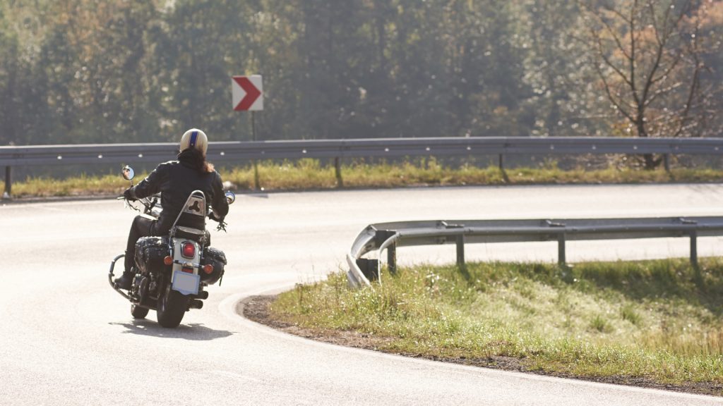 A person rides a motorcycle around a curve on a paved road with a metal guardrail and green foliage in the background.