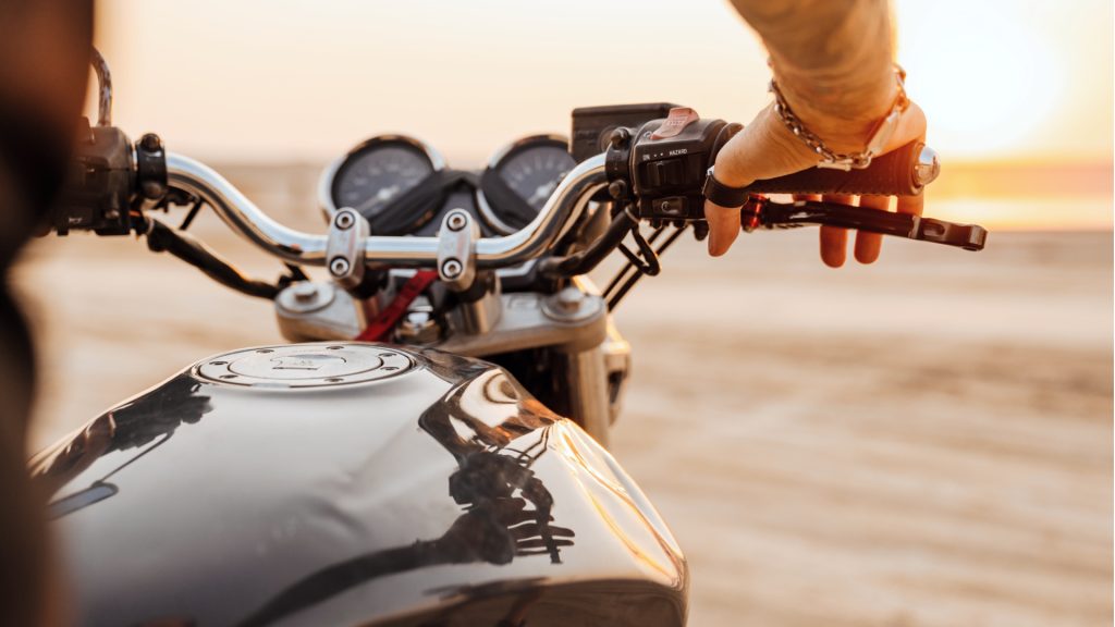 Close-up of a person’s hand gripping the handlebar of a motorcycle, with a blurred beach and sunset in the background.