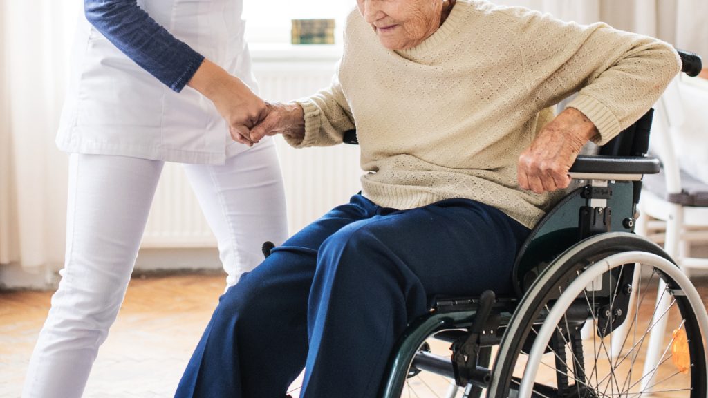 Un trabajador sanitario asiste a una persona mayor en silla de ruedas, sosteniendo su mano en una habitación bien iluminada.