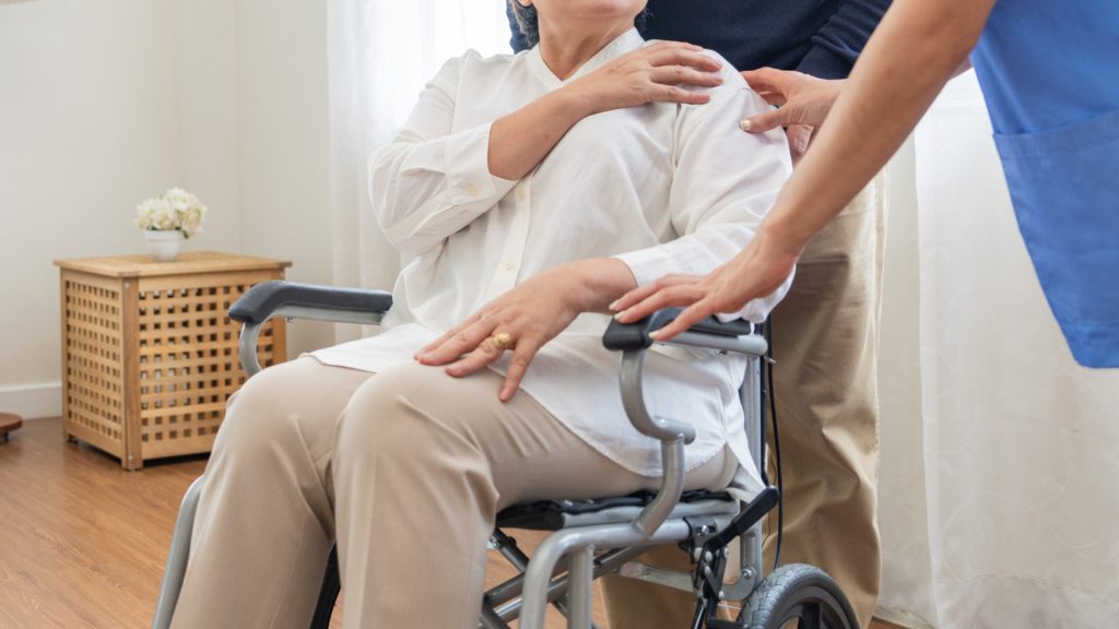 Una mujer mayor está sentada en una silla de ruedas con una mano sobre su hombro, asistida por dos personas en una habitación bien iluminada.