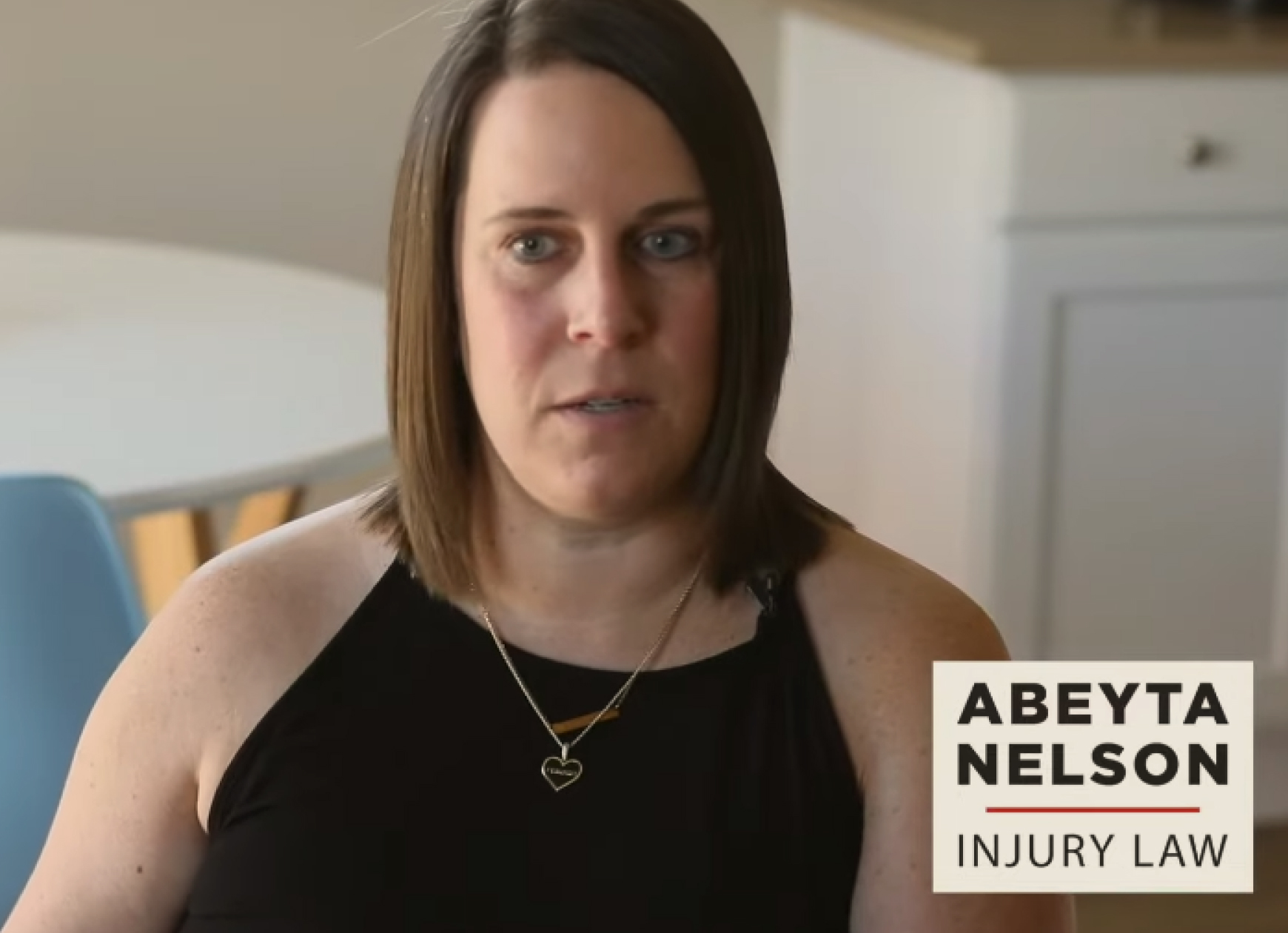 A woman with shoulder-length brown hair sits indoors, wearing a black sleeveless top and heart necklace; "Abeyta Nelson Injury Law" logo is visible in the lower right corner.