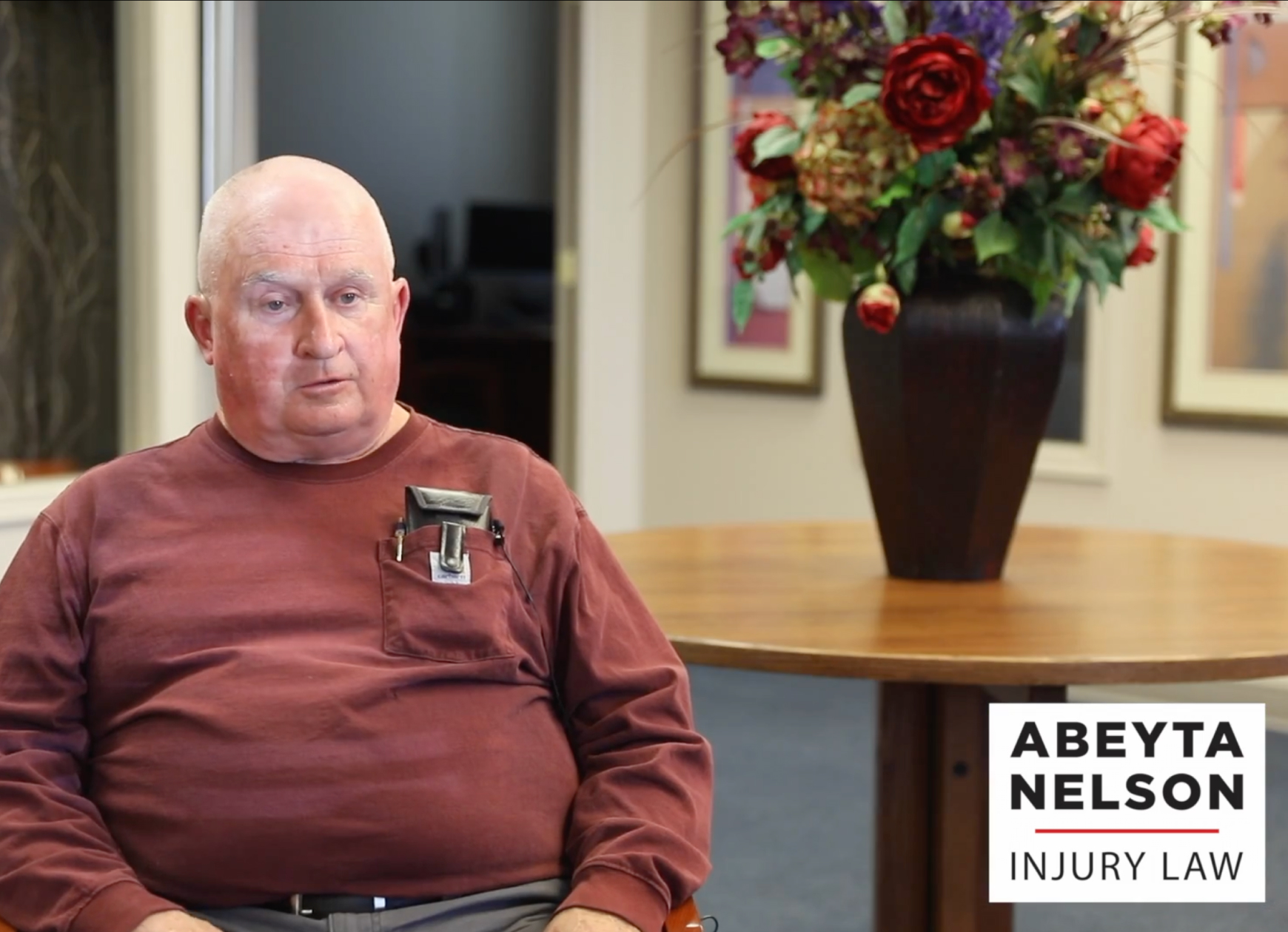 An older man in a maroon shirt sits in an office near a table with a vase of flowers. The Abeyta Nelson Injury Law logo is visible in the bottom right corner.