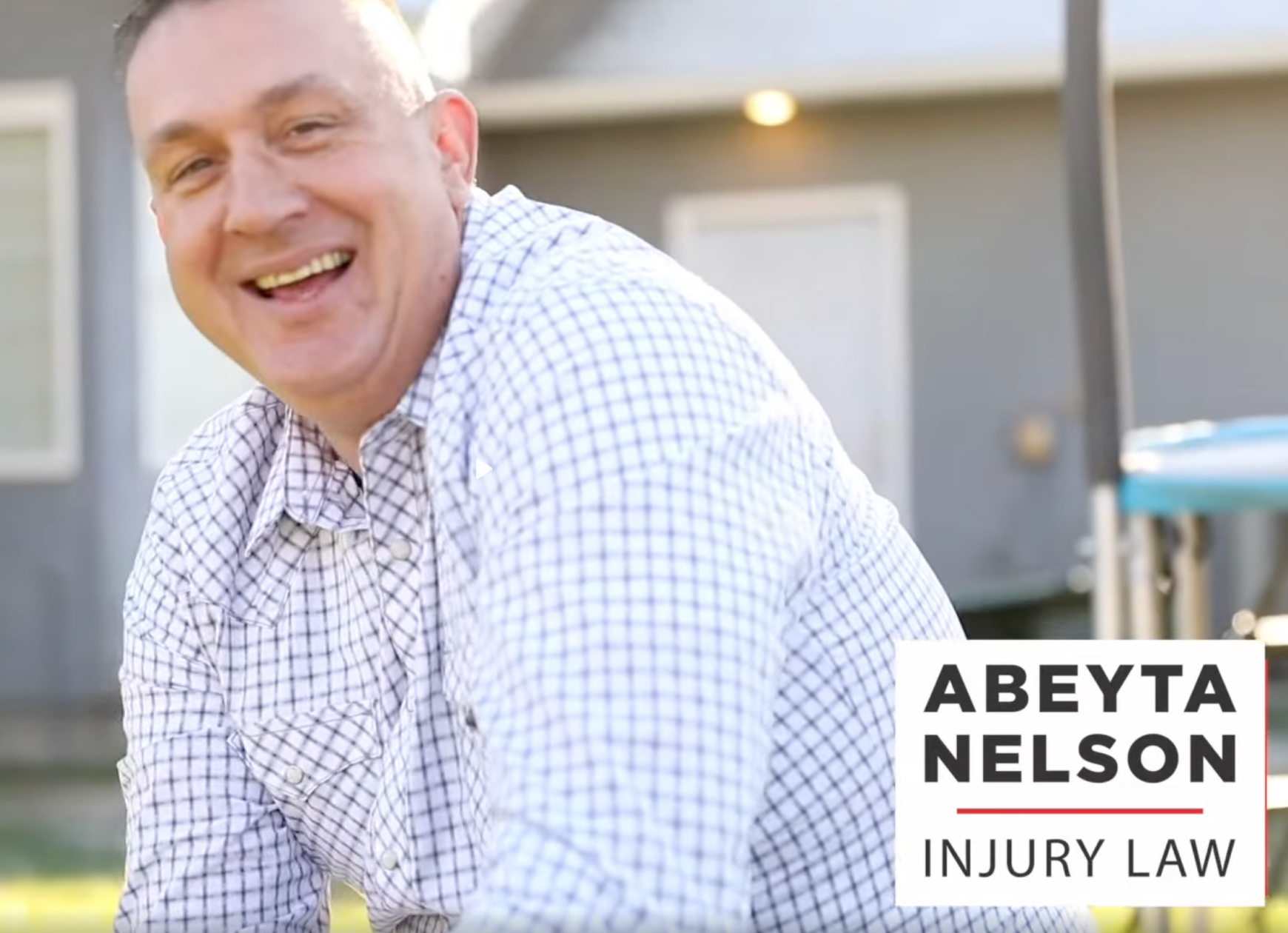 Smiling man in a checkered shirt sits outdoors with a house and playground in the background. "Abeyta Nelson Injury Law" logo appears in the lower right corner.