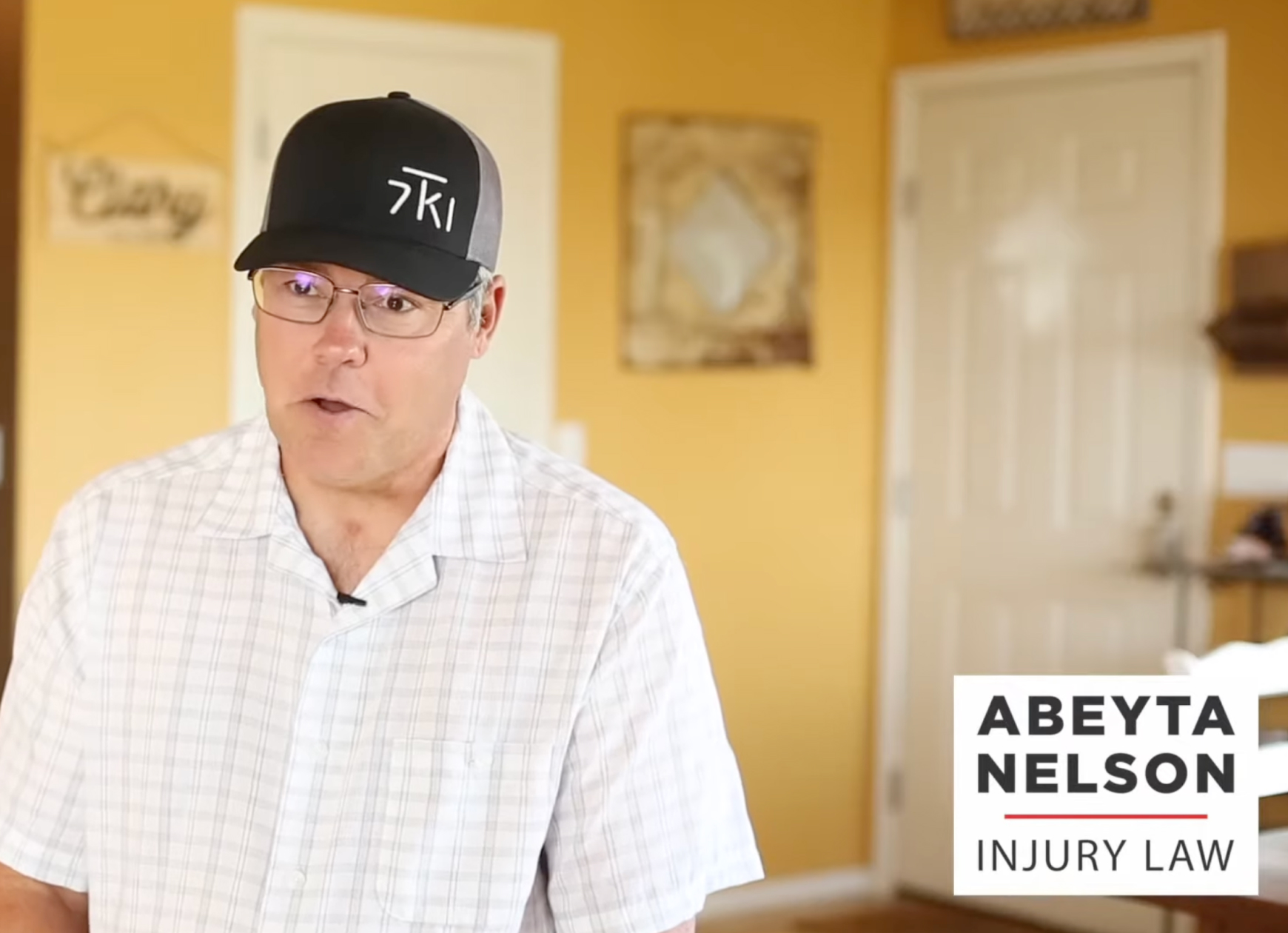 A man in a checkered shirt and black cap sits indoors; text in the lower right corner reads, "Abeyta Nelson Injury Law.