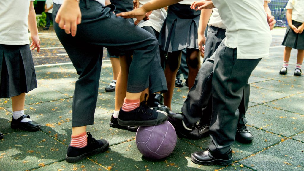 Children wearing school uniforms crowd around and play with a purple ball outdoors on a playground.