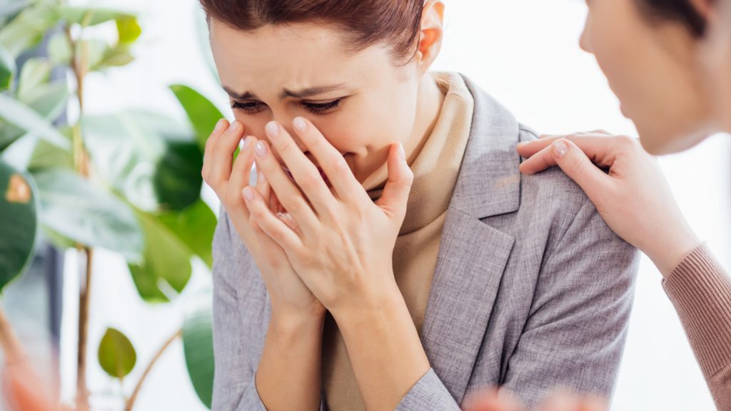 A woman in a gray blazer covers her face with her hands and appears to be crying, while another person comforts her with a hand on her shoulder.