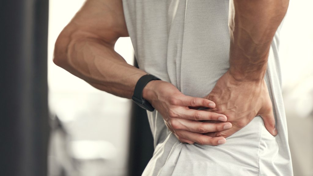 A person in a light gray shirt holds their lower back with both hands, suggesting discomfort or pain.