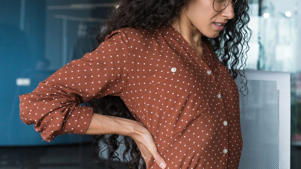 A person with long curly hair and glasses, wearing a brown polka dot shirt, holds their lower back and appears to be experiencing discomfort.