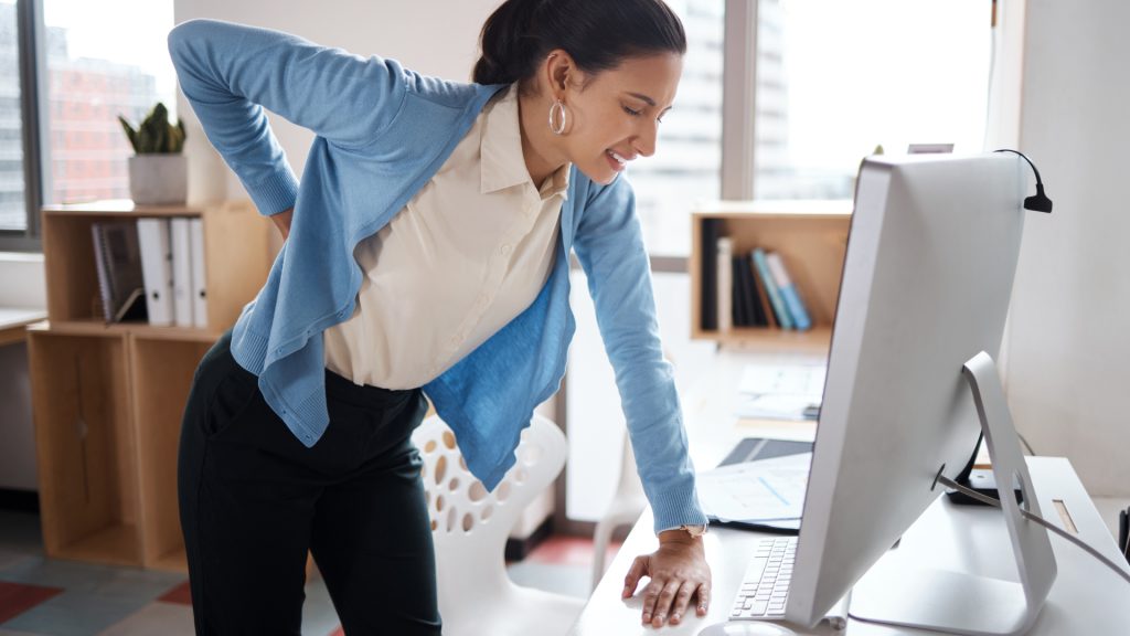 Woman standing at a desk, supporting herself with one hand on the table and holding her lower back with the other, appearing to experience back pain while working at a computer.
