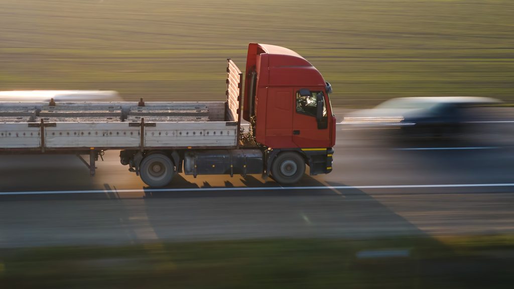 A red flatbed truck carrying concrete beams drives on a highway beside a blurred car, with grassy fields in the background.