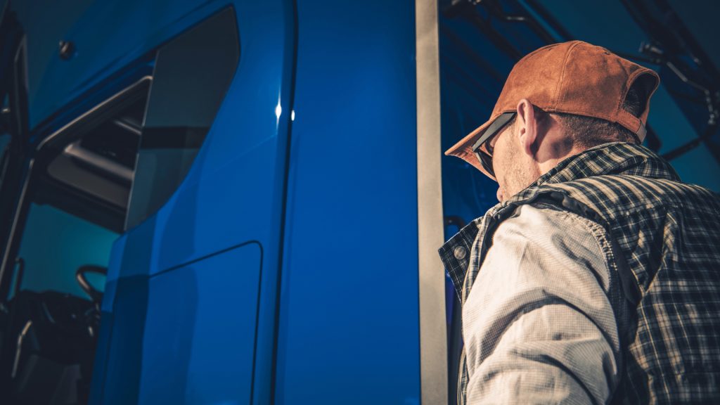 A man wearing a plaid shirt, vest, and brown cap stands next to a blue truck, looking toward the cab.