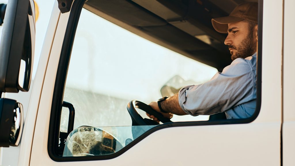A man wearing a cap drives a large vehicle, seen through the side window in daylight.