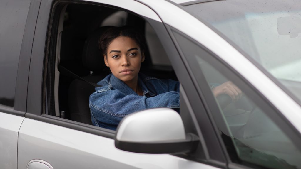A person with a neutral expression sits in the driver's seat of a car, looking out of the open window.