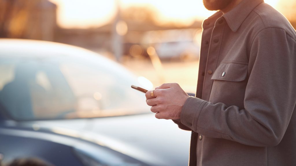 A person standing outdoors beside a car, using a smartphone, with the sun setting in the background.