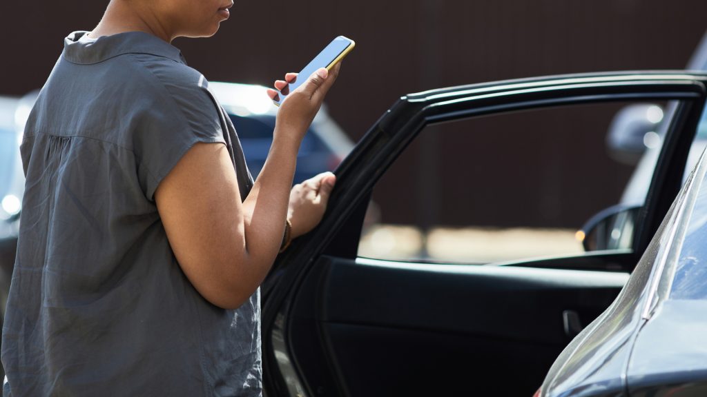 Person standing outside an open car door, holding a smartphone and speaking into it, with parked cars visible in the background.