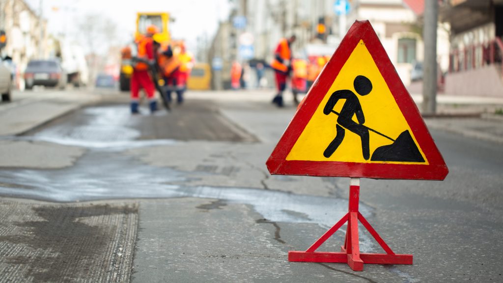 Una señal triangular de advertencia de obras viales está colocada en una calle con trabajadores con equipo de seguridad reparando la carretera al fondo.