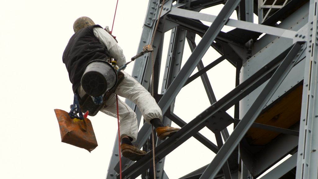 Un trabajador con equipo de seguridad está suspendido por cuerdas mientras realiza tareas de mantenimiento en una gran estructura metálica.