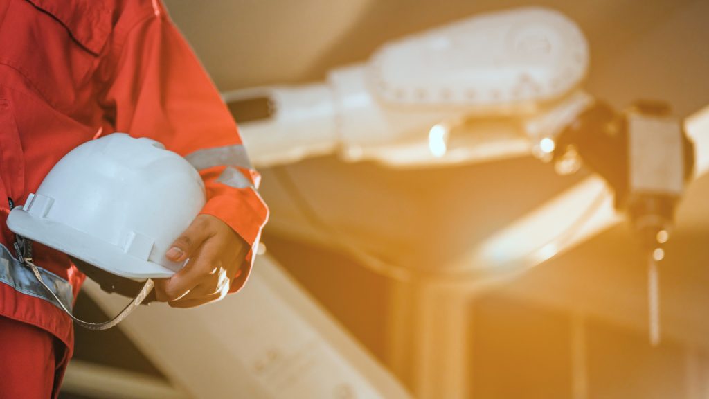 Una persona con un traje de seguridad rojo que sostiene un casco blanco está de pie cerca de un brazo robótico industrial en una fábrica.