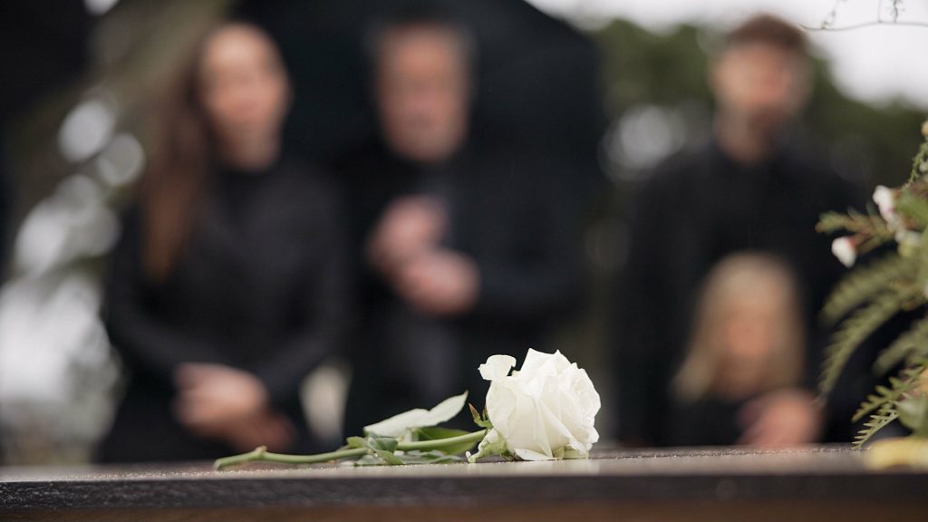 A white rose rests on a wooden surface in the foreground, with four blurred people dressed in black standing in the background, suggesting a funeral setting.