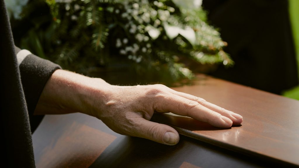 A person's hand rests on a wooden casket with a floral arrangement in the background.