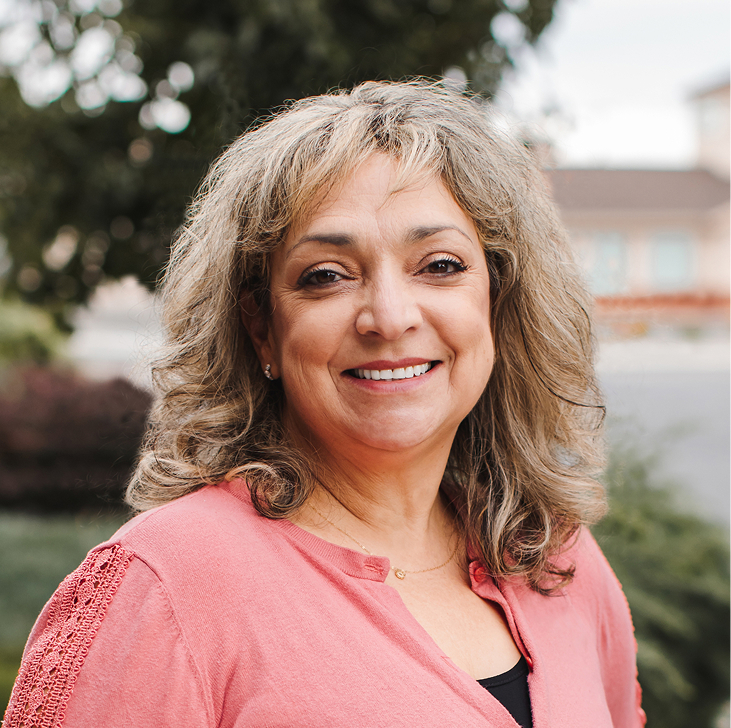 Una mujer con el pelo gris hasta los hombros y una blusa rosa sonríe a la cámara al aire libre, con vegetación y edificios al fondo.