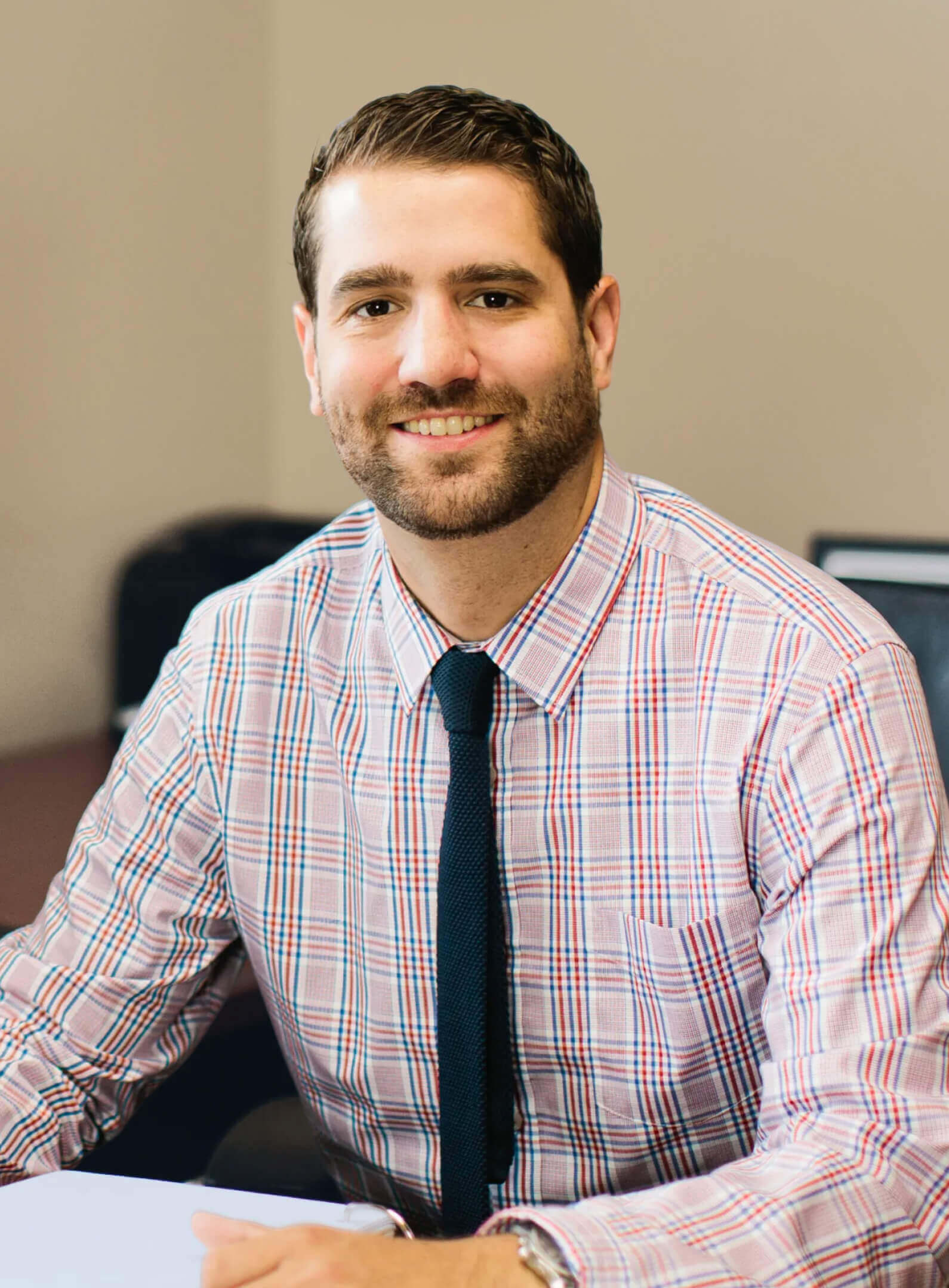 A man with short brown hair and a beard, wearing a plaid shirt and dark tie, sits at a desk and smiles at the camera.